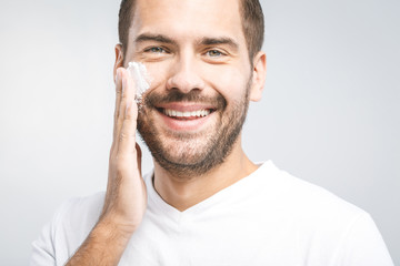 Skin care. Handsome young shirtless man applying cream at his face and looking at himself with smile while standing over gray background and looking at camera. Close-Up. Space for text.