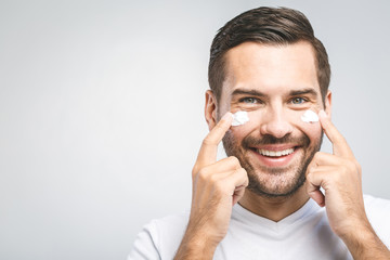 Obraz premium Skin care. Handsome young shirtless man applying cream at his face and looking at himself with smile while standing over gray background and looking at camera. Close-Up. Space for text.