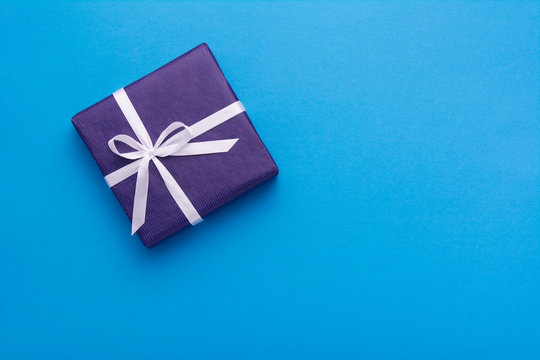 Hands Holding Wrapped Gift Box With Colored Ribbon As A Present For Christmas, New Year, Mother's Day, Anniversary, Birthday, Party,  On Blue Background, Top View. Present For A Colleague At Work.