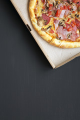 Top view, pizza with bacon, tomatoes, salami, cheese and mushrooms in a cardboard box on dark background. Copy space. From above, flat lay.