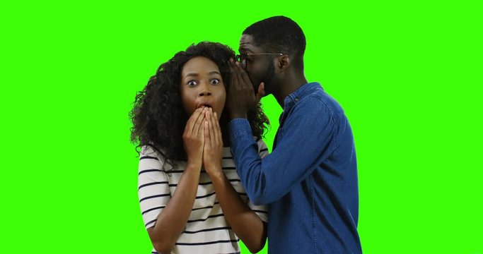 Nice young African American couple. Man in glasses whispering on the ear of beautiful surprised woman on the green screen background. Chroma key