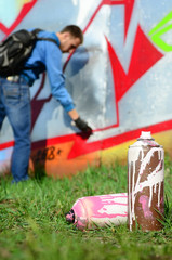 A few used paint cans against the background of the space with the wall on which the young guy draws a large graffiti drawing. Modern art of drawing walls in graffiti