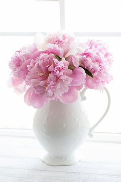 High key photograph of a bouquet of large pink peonies in a white pitcher in the window sill