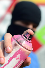 A young graffiti artist in a blue jacket and black mask is holding a can of paint in front of him against a background of colored graffiti drawing. Street art and vandalism concept