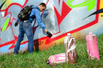 A few used paint cans against the background of the space with the wall on which the young guy draws a large graffiti drawing. Modern art of drawing walls in graffiti