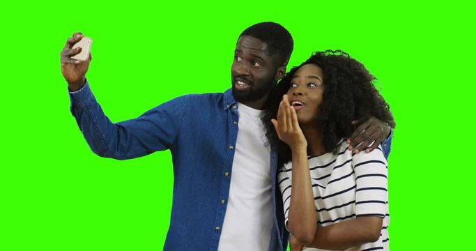 Happy young African American man and woman posing and making nice selfies on the smartphone in male hands on the green screen background. Chroma key.