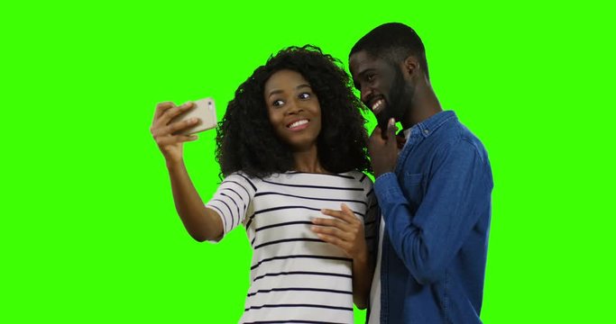 Joyful young African American couple posing funny and taking nice selfies on the smartphone in female hands on the green screen background. Chroma key.
