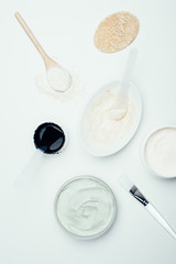 flat lay with sponge, various clay masks in containers, clay powder, spoons, plate and brush isolated on white surface