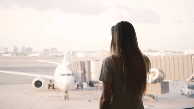 Young European girl talks on smartphone and smiles, ends call at airport terminal window, airplane in the background.