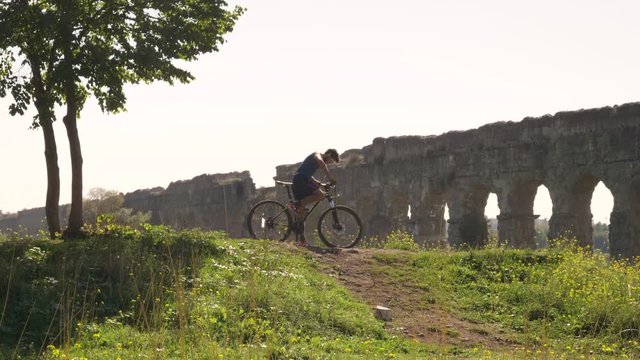 Cyclist riding bike uphill on dirt road in front of majestic ancient roman aqueduct. Young attractive athletic man with blue t-shirt and shorts sportswear in parco degli acquedotti in rome