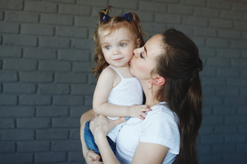 Mother and daughter in white T-shirts and blue jeans, hugging and smiling in the studio on a dark gray brick background.