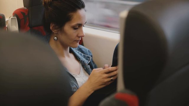 Casual female passenger on the train sits in a comfortable seat using smartphone e-commerce app, looking at the window.