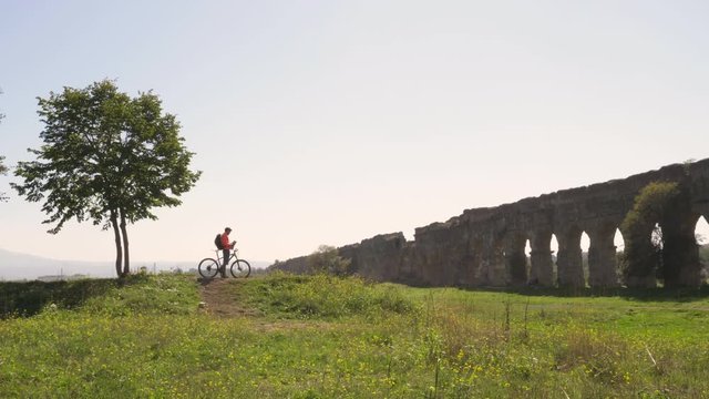 Cyclist with bike reads map for direction in front of ancient roman aqueduct in a park. Young attractive athletic man with orange sportswear and backpack in parco degli acquedotti in Rome under a tree
