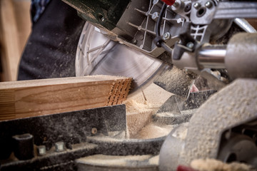 Close up of experienced carpenter in work clothes and small buiness owner working in woodwork workshop, using a circular saw to cut through a wooden plank, on the table is a hammer and many tools