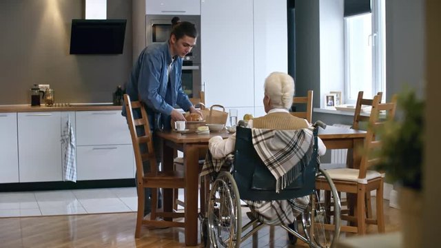 PAN of young male caregiver serving buns with butter and coffee to elderly woman in wheelchair