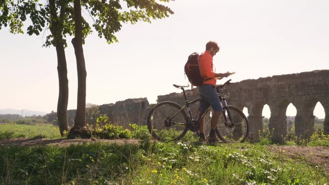 Cyclist with bike reads map for direction in front of ancient roman aqueduct in a park. Young attractive athletic man with orange sportswear and backpack in parco degli acquedotti in Rome under a tree