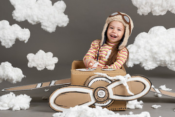 the child girl plays in an airplane made of cardboard box and dreams of becoming a pilot, clouds of cotton wool on a gray background