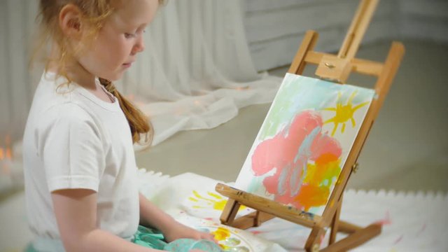 Beautiful red-haired girl sits on the floor in the studio in front of the easel and draws a brush and colored paints