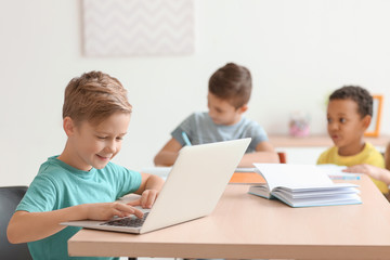 Cute boy with laptop doing homework in classroom at school