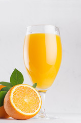 Tropical citrus yellow juice in elegance wineglass closeup with ripe oranges and green leaves on soft white wood background, vertical.
