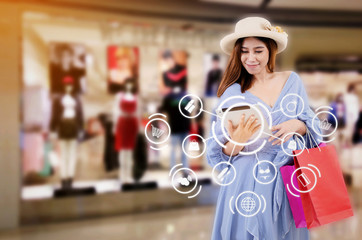 asian woman watching special offer in tablet and holding shopping bag while standing at new collection clothes shop in department store shopping mall with icon product diagram, shopping online concept