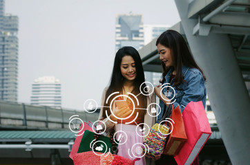 two beautiful asian young woman watching special offer in mobile phone and holding shopping bag in big city with icon product diagram, technology, summer sale, payment and shopping online concept