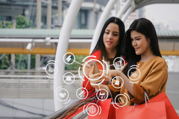 two beautiful asian young woman watching special offer in mobile phone and holding shopping bag in big city with icon product diagram, technology, summer sale, payment and shopping online concept