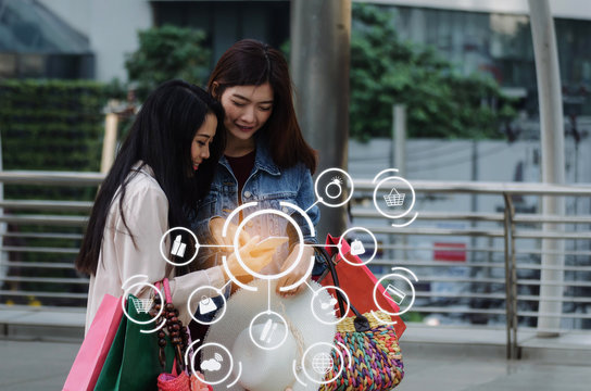 two beautiful asian young woman watching special offer in mobile phone and holding shopping bag in big city with icon product diagram, technology, summer sale, payment and shopping online concept