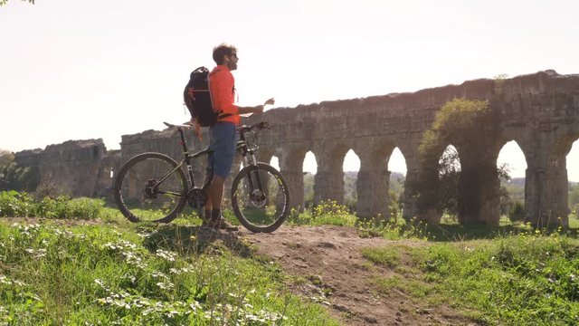 Cyclist with bike reads map for direction in front of ancient roman aqueduct in a park. Young attractive athletic man with orange sportswear and backpack in parco degli acquedotti in Rome under a tree