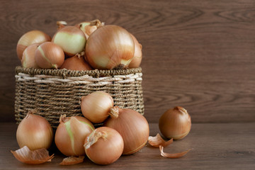 Many onion bulbs in the box. Large wicker basket with onions. Vegetables on the table next to the basket.