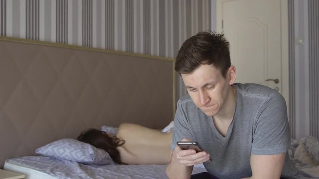 Man is sitting on the bed with a phone on the background of a naked woman.
