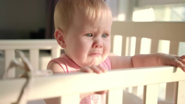 Sad baby crying in cot at home. Unhappy toddler standing in crib. Upset child in bed looking mom. Unhappy child wake up at morning. Sadness baby emotion. Child fear