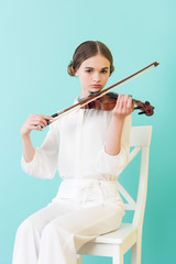 teen girl playing violin and sitting on chair, isolated on turquoise