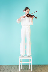 teen girl in white outfit playing violin and standing on chair, on turquoise