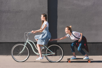 side view of woman riding bicycle and towing her smiling female friend on skateboard