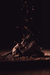 close up shot of grated dark chocolate falling on pieces of chocolate on black background - obrazy, fototapety, plakaty
