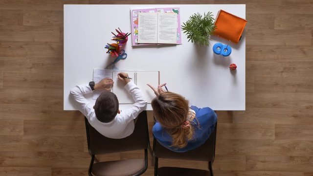Young mother helping son with homework, sitting behind table, kid doing homework top shot parquet wooden floor