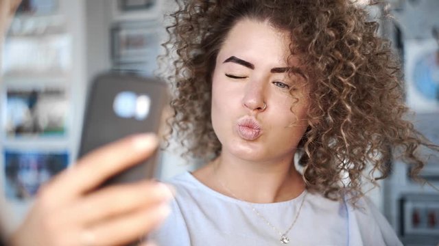 Video portrait like kinky beautiful young woman smiles to the camera and makes selfie with her phone in different poses, girl makes photos to the social networks, photo after making hairstyle