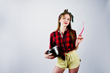 Obraz premium Young funny housewife in checkered shirt and yellow shorts pin up style with saucepan and kitchen spoon isolated on white background.