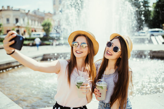 Beautiful female friends pose at camera of modern smart phone, make selfie, sit together in outdoor at street drink shake, have positive expressions. Pleased women colleagues meet after work for lunch - Powered by Adobe