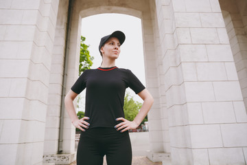 Young concentrated athletic beautiful brunette girl in black uniform and cap doing sport exercises, warm-up before running, standing in city park outdoors. Fitness, healthy lifestyle concept.