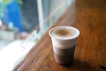 A plastic glass of chocolate milkshake with layer of cream cheese, topped with cocoa powder on wooden table, Relax on weekend, Summer drink concept.