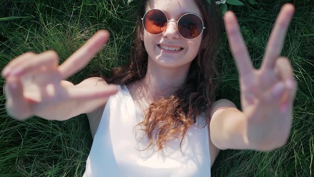 SLOW MOTION: Top view shot of a Caucasian girl in a white floral dress lying in grass and shows a sign of peace