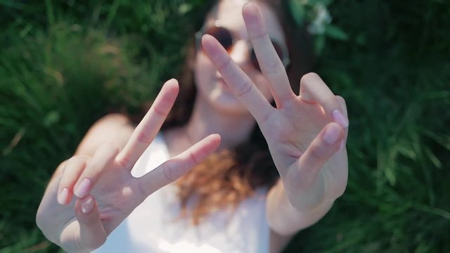 SLOW MOTION: Top view shot of a Caucasian girl in a white floral dress lying in grass and shows a sign of peace