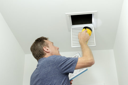 Checking Air Ducts in Home HVAC System. Man inspecting air ducts shining a flashlight through a small square ceiling vent into ducting pipes. Mature male examining the condition of air ducts at home.