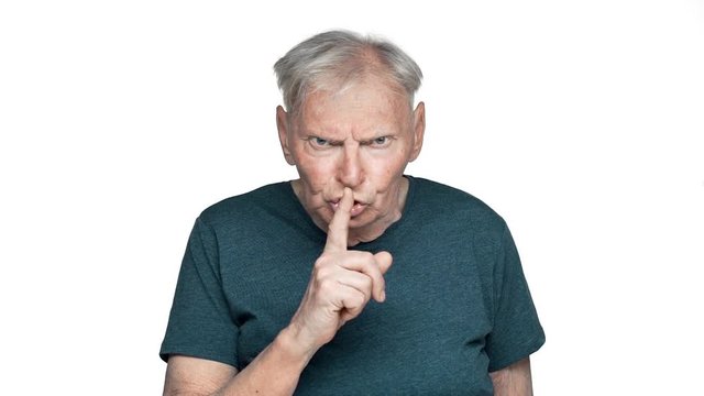 Portrait of angry old aged man 80s in basic t-shirt posing with strict gaze and asking keep silence putting index finger on lips slow motion, isolated over white background