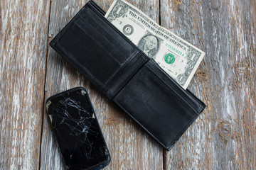 Crashed black smartphone is lying in an empty black leather wallet. One dollar in the purse. No money to buy a new phone. Poverty and unemployment. Time to take a loan for electronics. Wooden table.