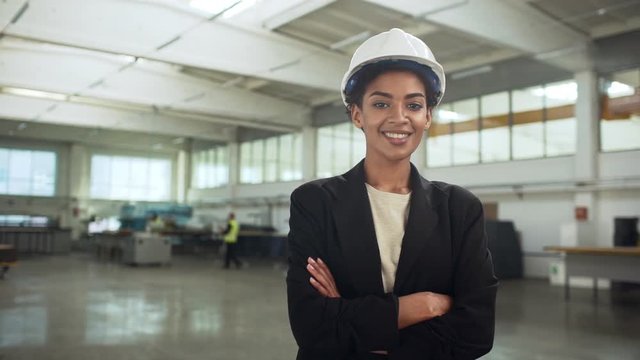 Portrait of successful african american woman in helmet and suit standing with arms crossed in big bright room of manufacture slow motion