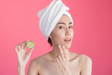 beautiful asia woman with towel on head with perfect clean skin smiling holding cucumber slices over pink background. Beauty cosmetology and spa.