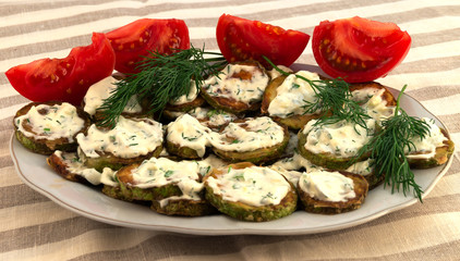 Snack from fried zucchini slices with sour cream and dill, next to several slices of tomato.